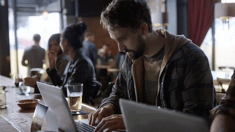 Man working on his laptop at a bar/coffee shop looks to his left to see a row of people that look exactly like him on their laptops too
